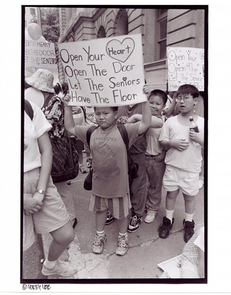 Image of young An Rong Xu, holding up a sign that says "Open Your Heart, Open the Door, Let the Seniors Have the Floor". Photographed by Corky Lee, courtesy of An Rong Xu.