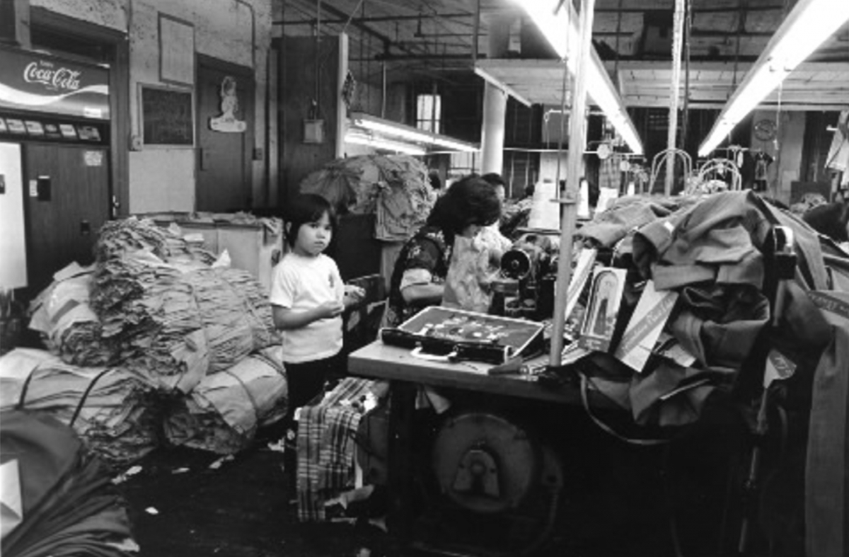 Interior of a Chinatown sewing factory. A young girl stands beside two women working. Heaps of fabric surround them.