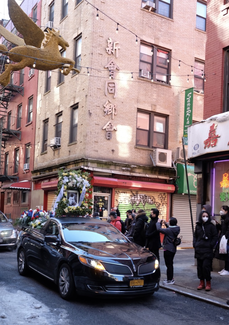 Photo of black sedan with funeral wreath going through New York Chinatown with people watching on the sidewalk.
