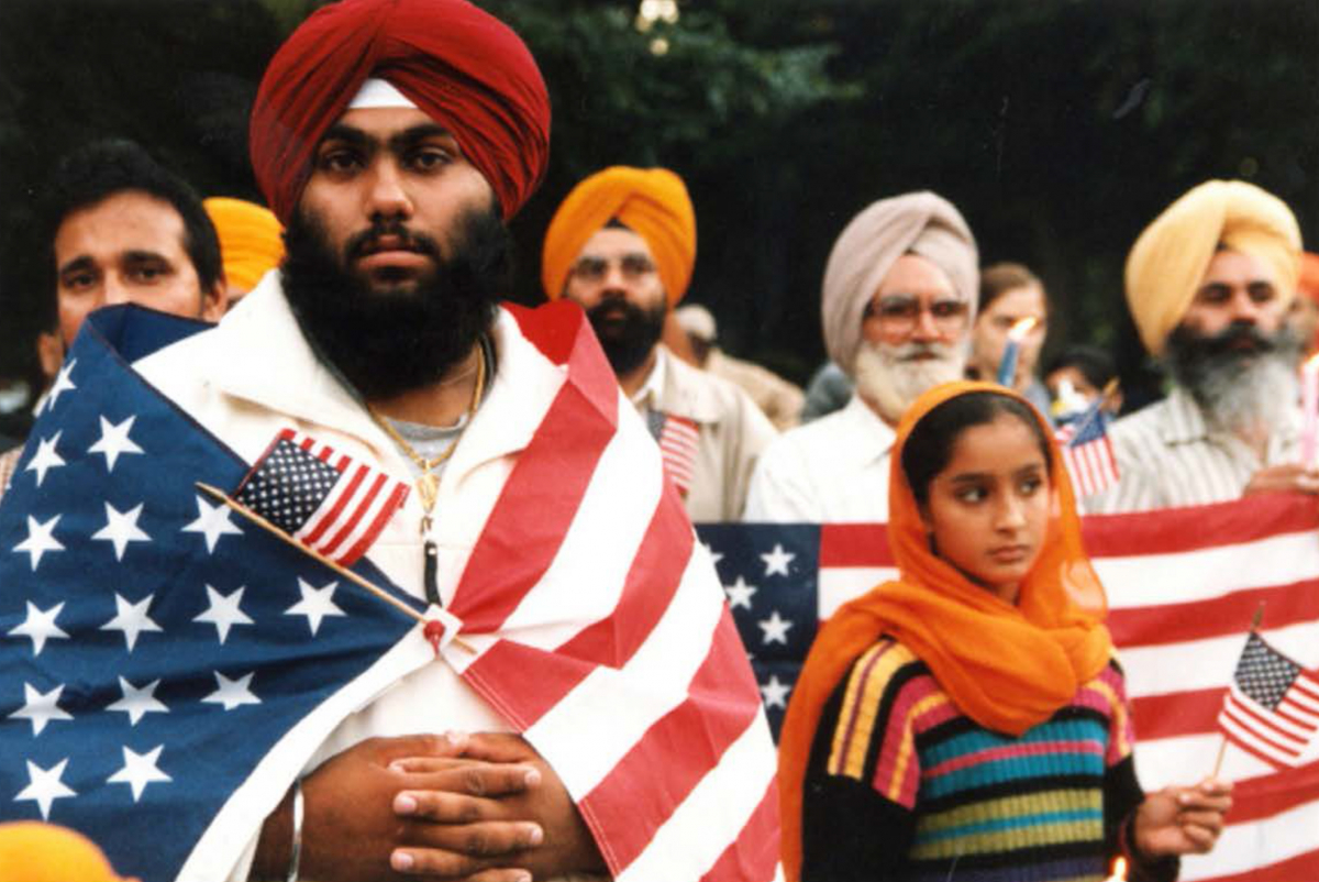 A man with red turban has an American flag draped around his shoulders. A girl with orange headscarf stands besides him wearing a colorful striped sweater and other Sikhs of all ages are in the background.