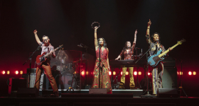 A production shot of five members of a band, their arms raised and pointed to the sky