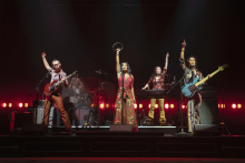 A production shot of five members of a band, their arms raised and pointed to the sky